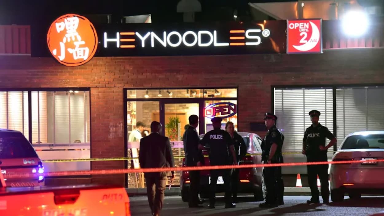 Photo de policiers se tenant debout à l'extérieur d'un restaurant le soir. Sur l'enseigne, on peut lire « HeyNoodles ».