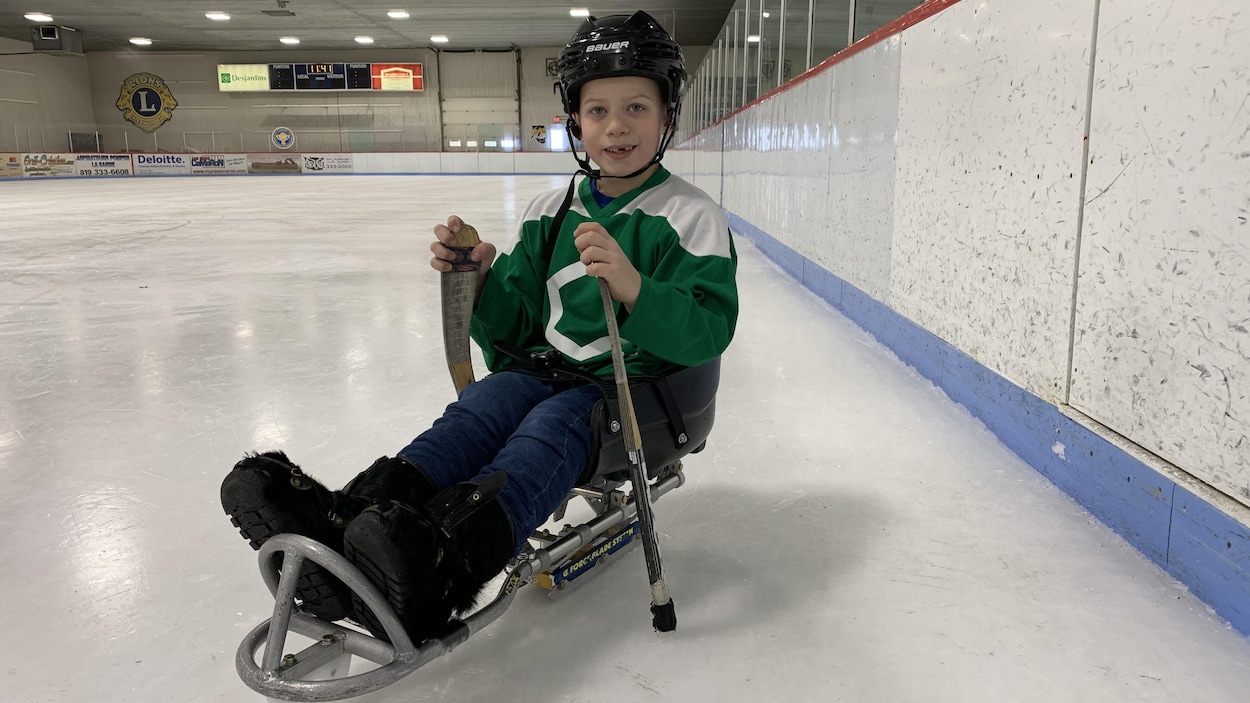 Tyler est assis avec les jambes devant lui sur une petite luge sur une patinoire dans un aréna. Il tient deux petits ba^tons de hockey dans ses mains.