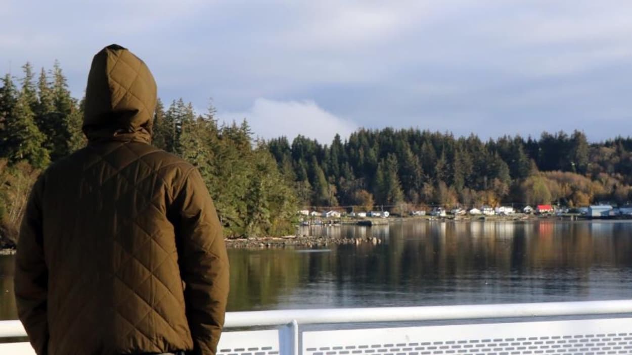 Une personne debout sur le pont d'un traversier regarde une côte où se trouvent plusieurs maisons adossées à une forêt dense.
