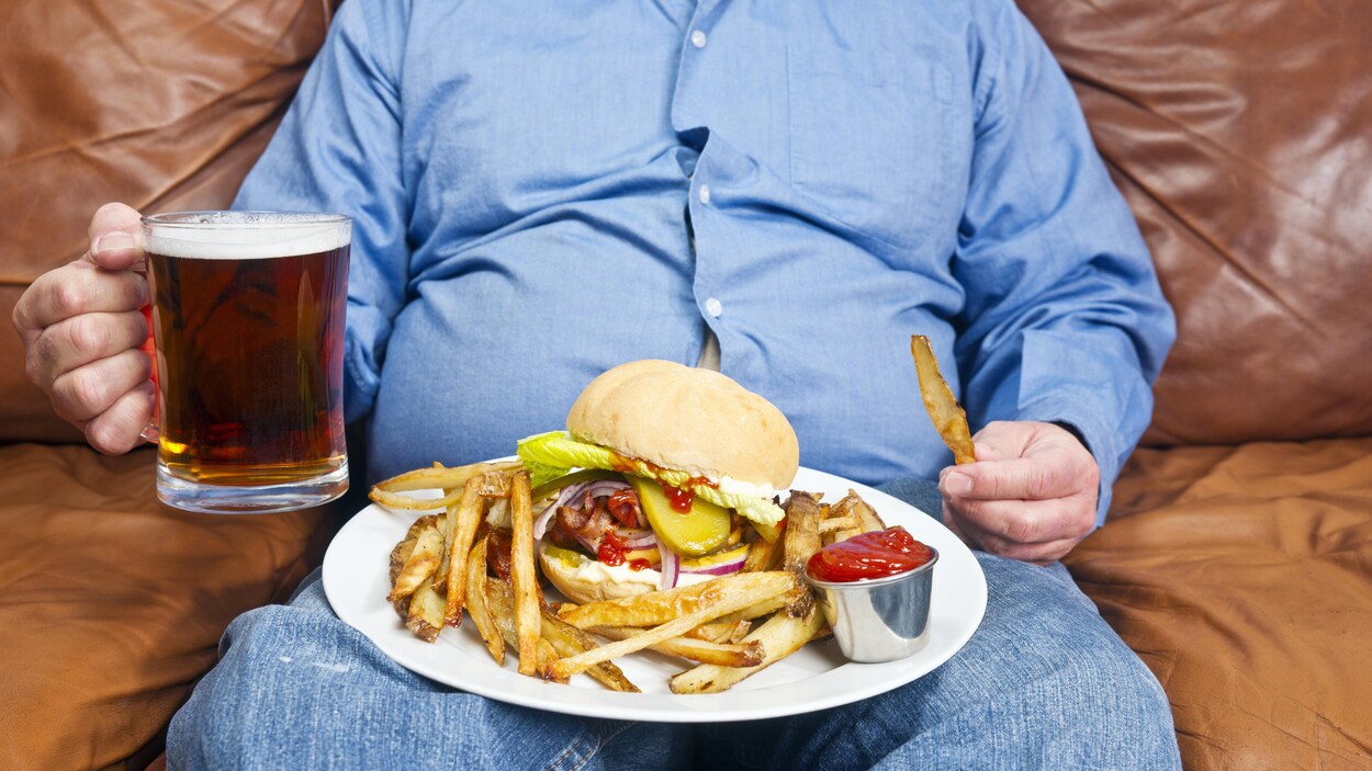 Un homme en surpoids boit une bière et mange un hamburger et des frites. Un homme en surpoids boit une bière et mange un hamburger et des frites.