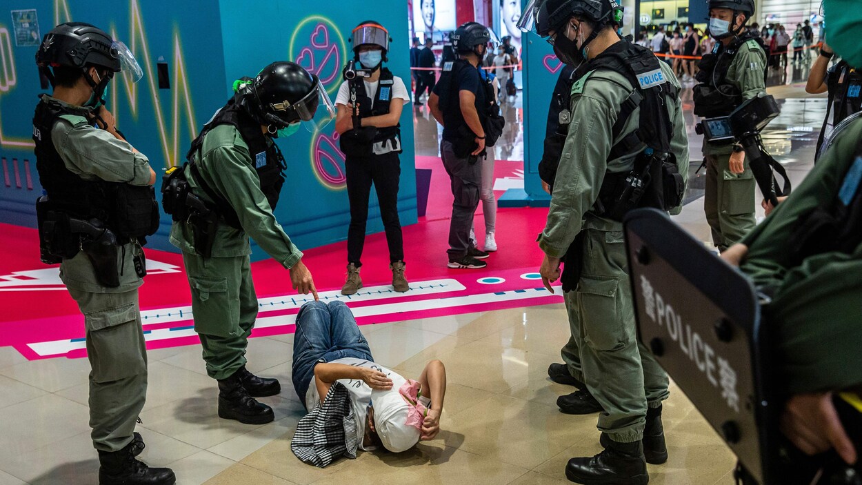 Des policiers entourent une femme qui est couchée par terre sur le dos. L'un d'entre eux pointe le doigt dans sa direction.
