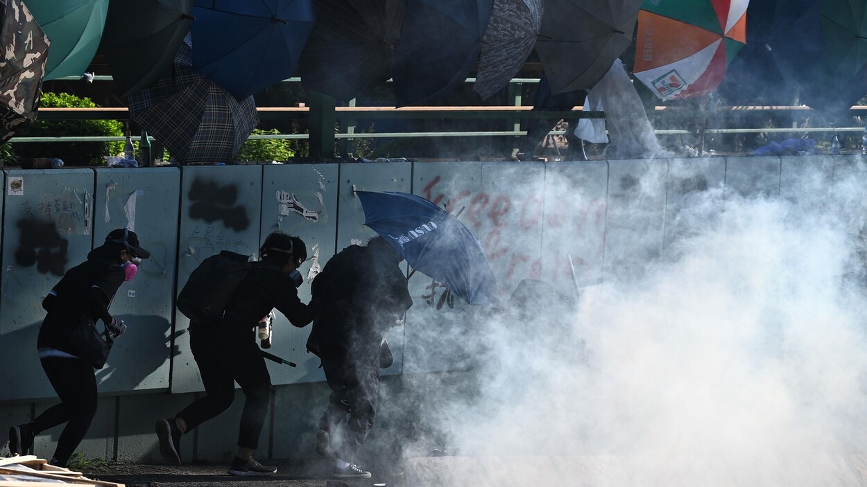 Trois manifestants masqués marchent derrière un nuage de gaz lacrymogène.