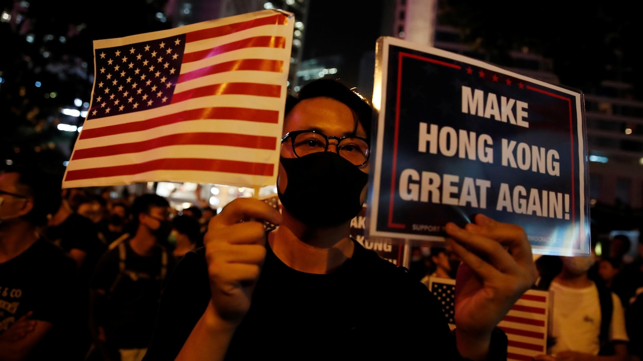 Un homme avec un masque tient un petit drapeau américain et une pancarte où est écrit : « Make Hong Kong great again! »