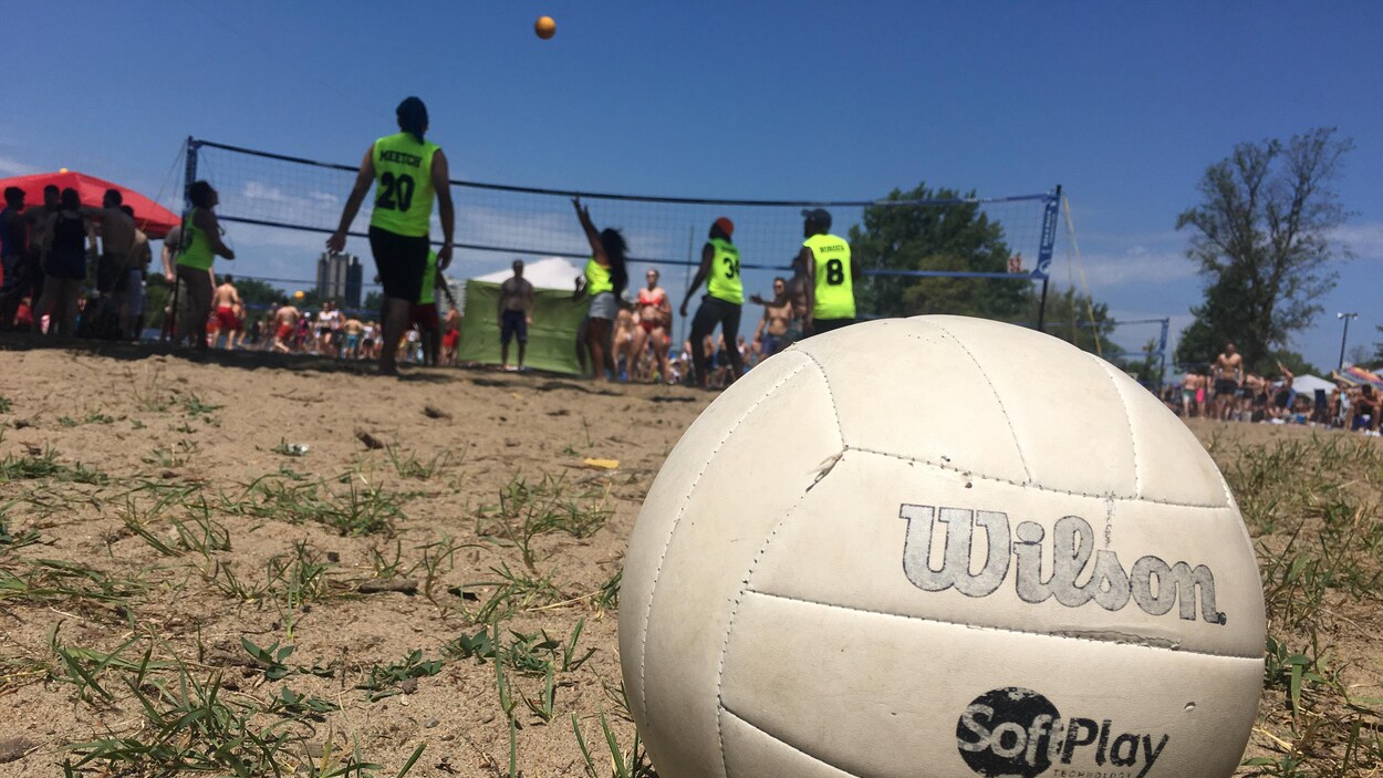 Un ballon de volleyball en avant-plan. Derrière, des gens jouent au volleyball de plage.