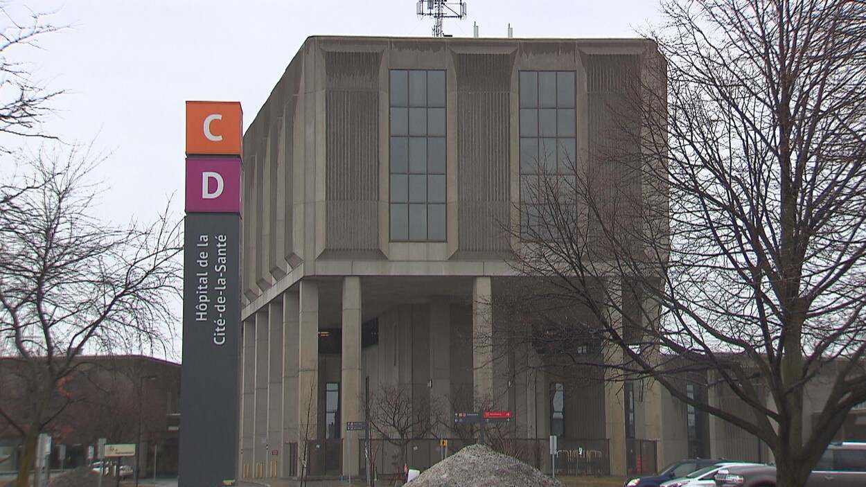 Un panneau portant le nom de l'hôpital à la verticale devant un bâtiment en béton par une journée pluvieuse de printemps.
