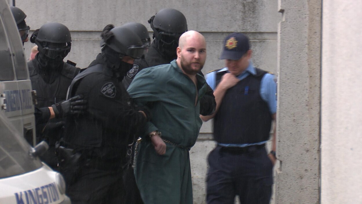 Photo d'un homme menotté habillé en vert qui est escorté par des policiers vêtus de noir. Photo d'un homme menotté habillé en vert qui est escorté par des policiers vêtus de noir.
