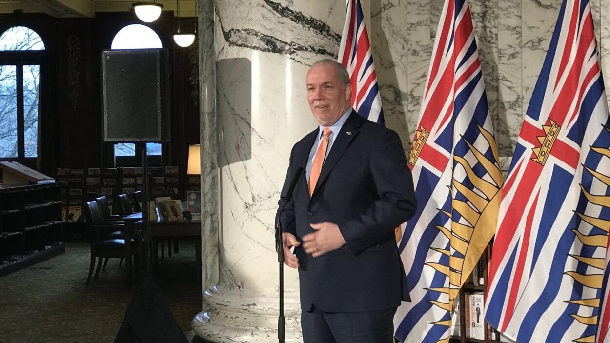John Horgan s'approche d'un microphone installé devant trois drapeaux de la province. John Horgan s'approche d'un microphone installé devant trois drapeaux de la province.