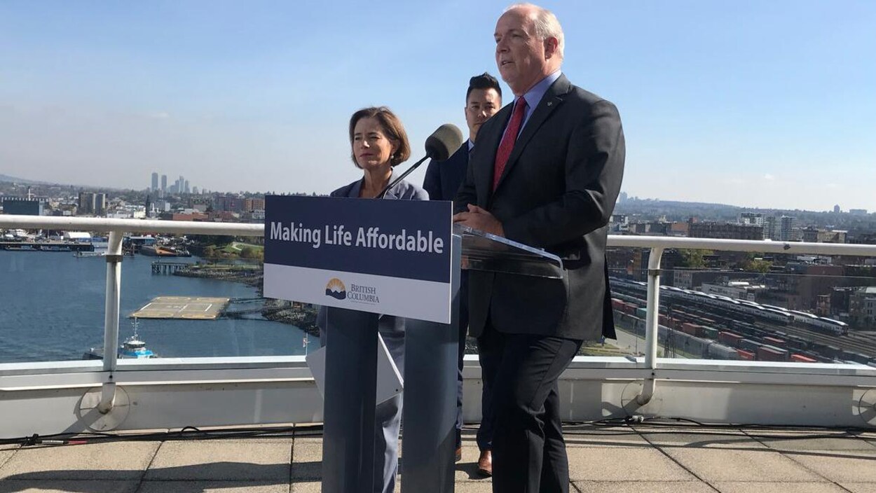 John Horgan debout devant un lutrin et un micro, avec une femme et un homme derrière lui, avec en arrière-plan de l'eau et des rails de chemin de fer.