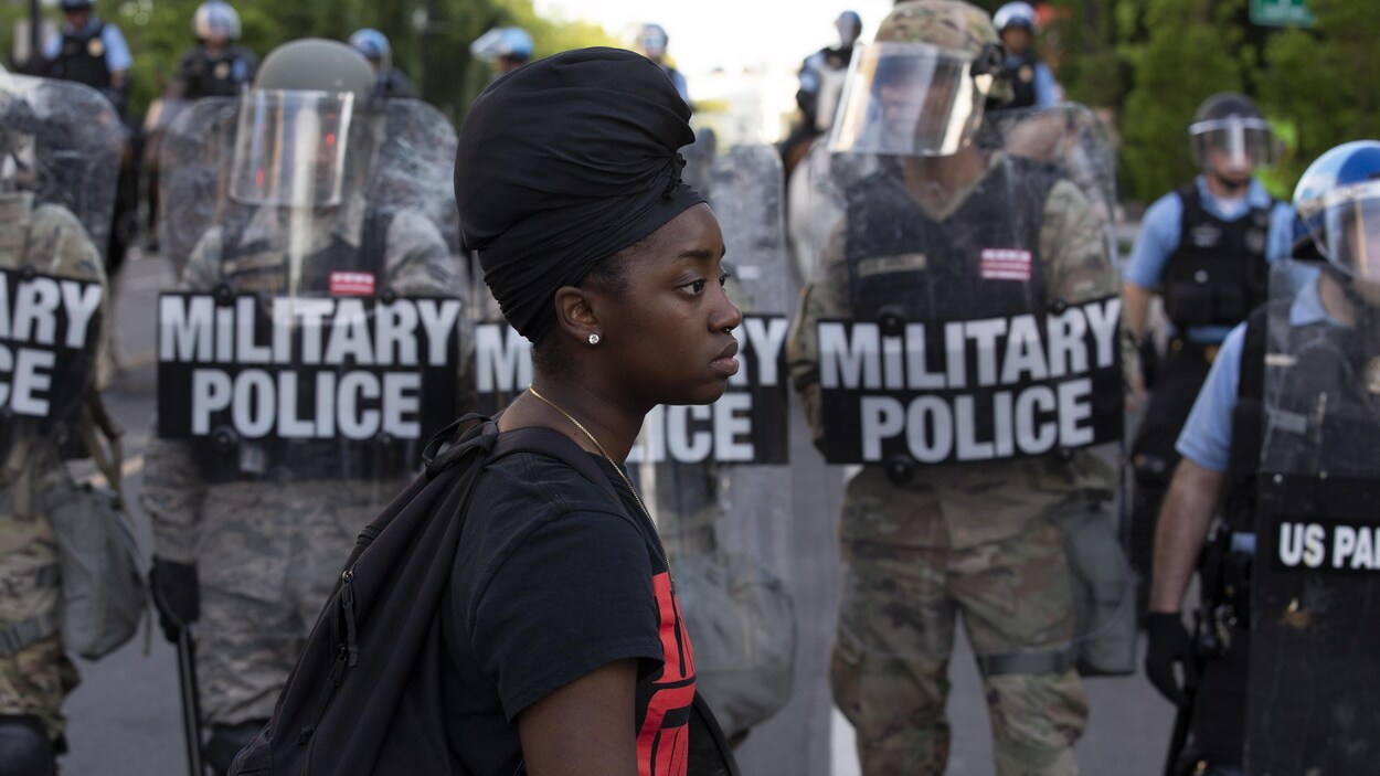 Une jeune femme noire de profil marche devant des policiers en tenue antiémeute portant la mention « police militaire ».
