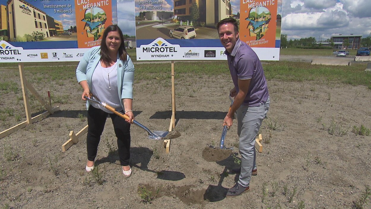 Julie Morin, mairesse de Lac-Mégantic, et Mathieu Mault, président de Activar avec des pelles remplies de terre devant une affiche qui annonce le nouveau complexe hôtelier de Lac-Mégantic.