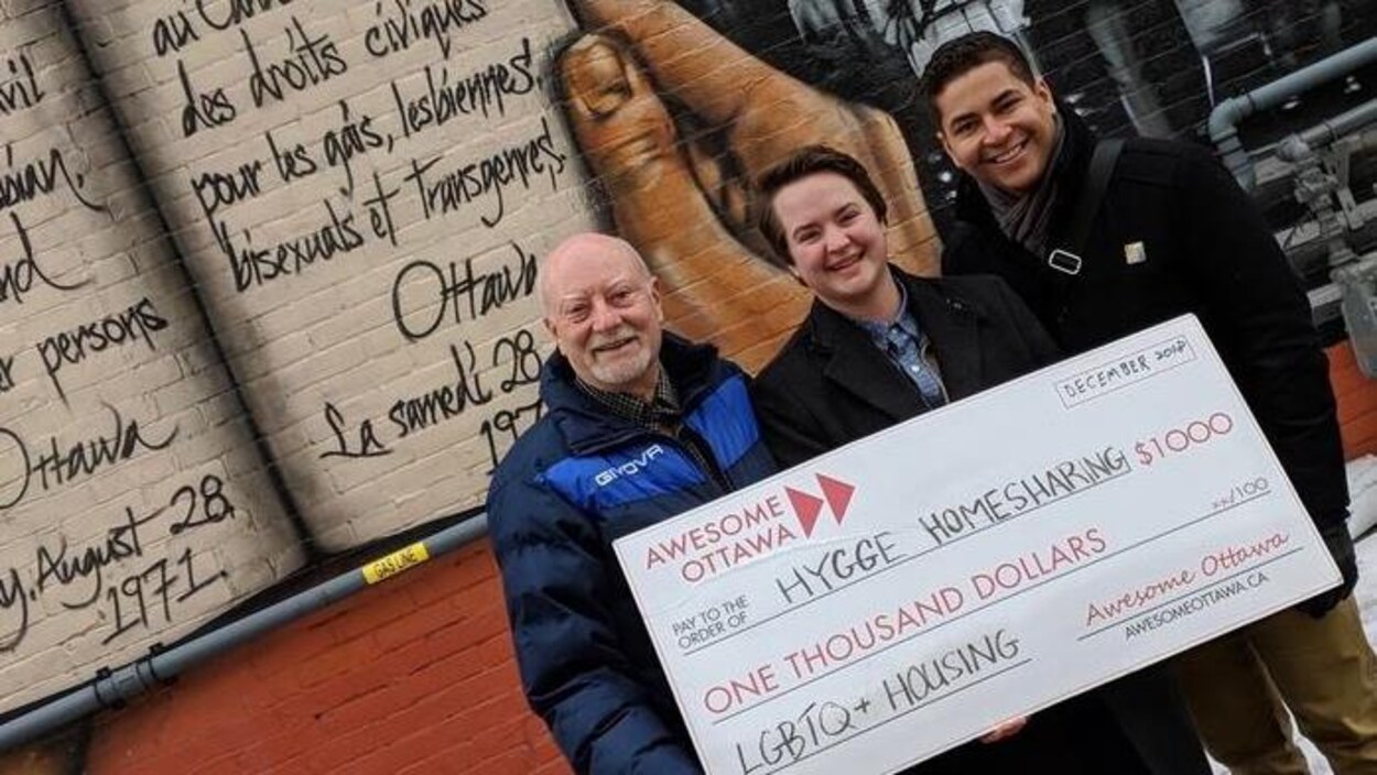 Deux hommes et une femme posent pour la caméra en tenant un gros ch`que devant une murale peinte sur le mur extérieur d'un bâtiment.
