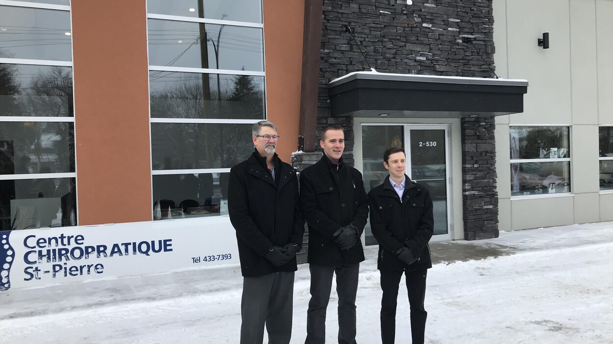 Trois hommes debout côte à côte devant un bâtiment neuf par une journée d'hiver.