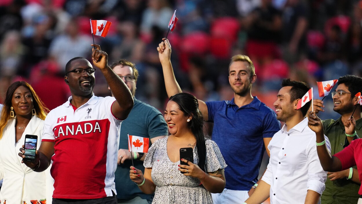 Une douzaine de personnes de diverses origines ethniques sont debout en rang sur un terrain de football à la mi-temps. Ils agitent de petits drapeaux du Canada et regardent autour d'eau. Un homme au centre porte un chandail rouge et blanc décoré du mot « Canada ».
