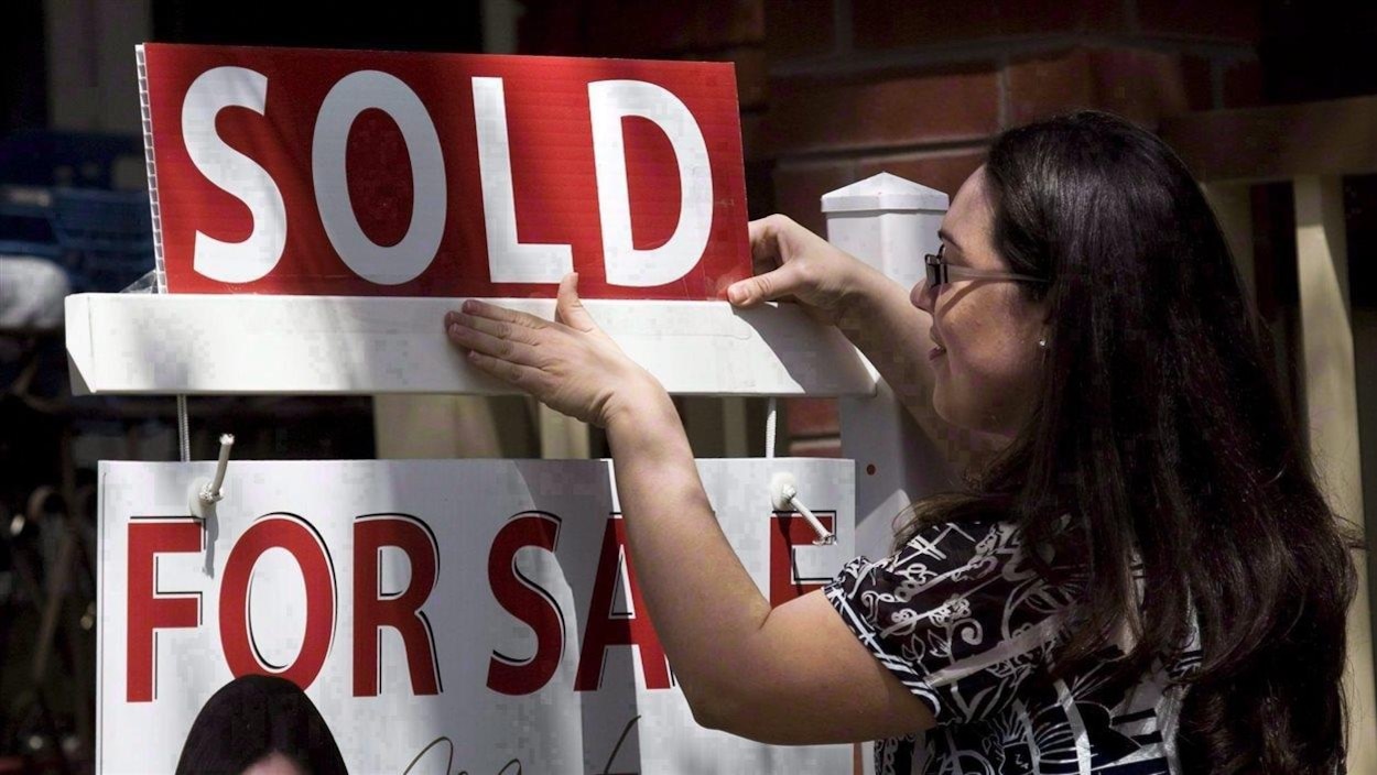 Une femme brune accroche un panneau SOLD (vendu) au-dessus d'un panneau à vendre en lettres rouges et blanches.