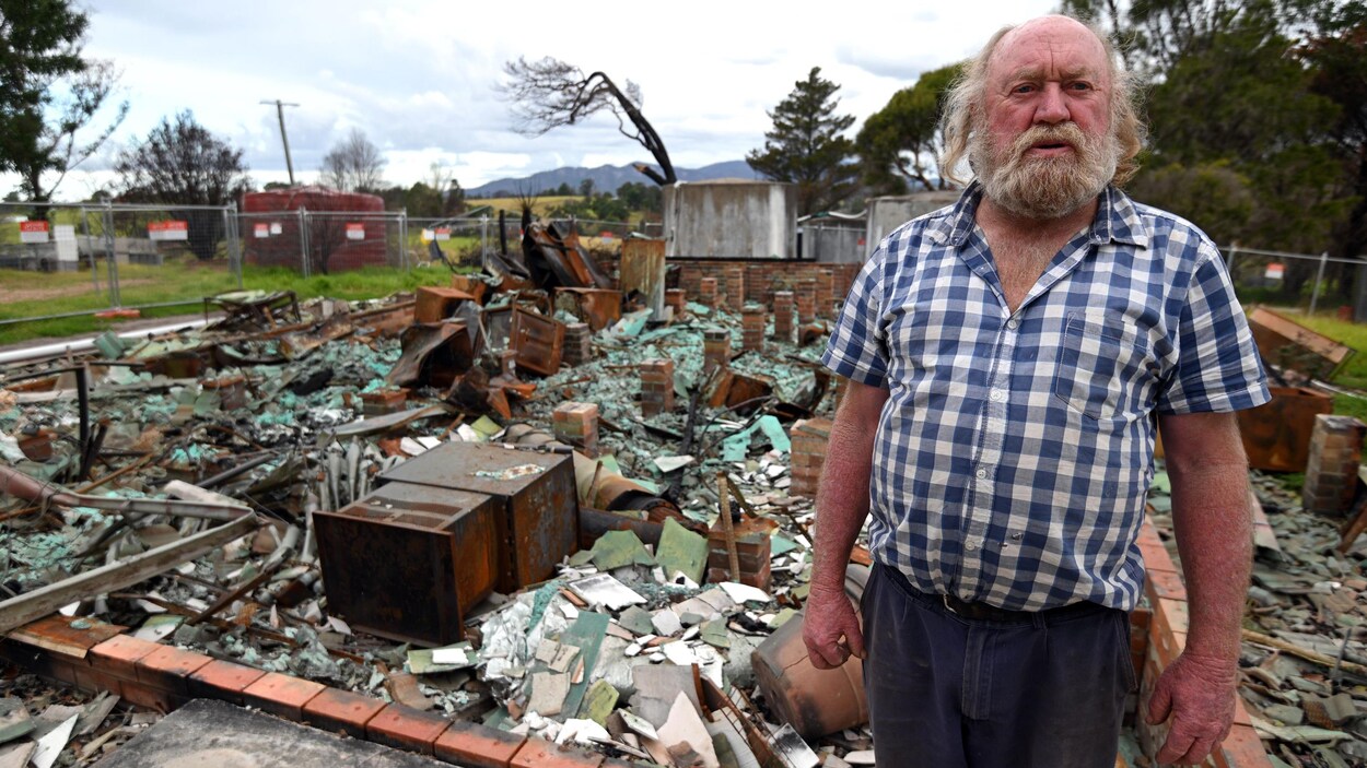 Un homme âgé se tient debout devant les ruines de sa maison.