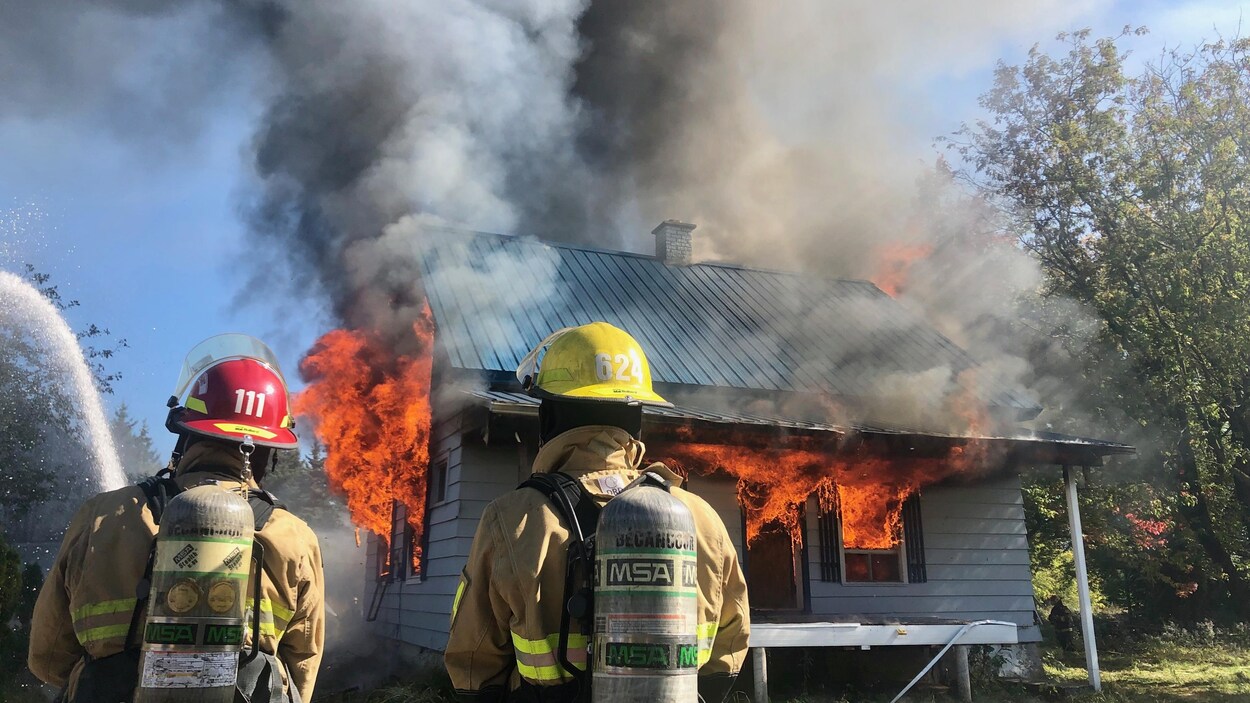 Deux pompiers regardent une petite maison qui brûle.