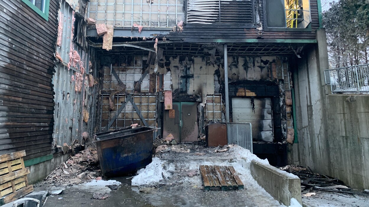 L'espace de livraison du IGA a été touché par l'incendie. Des bouts du mur extérieur sont brûlés. Un conteneur de déchets est au centre de la photo.