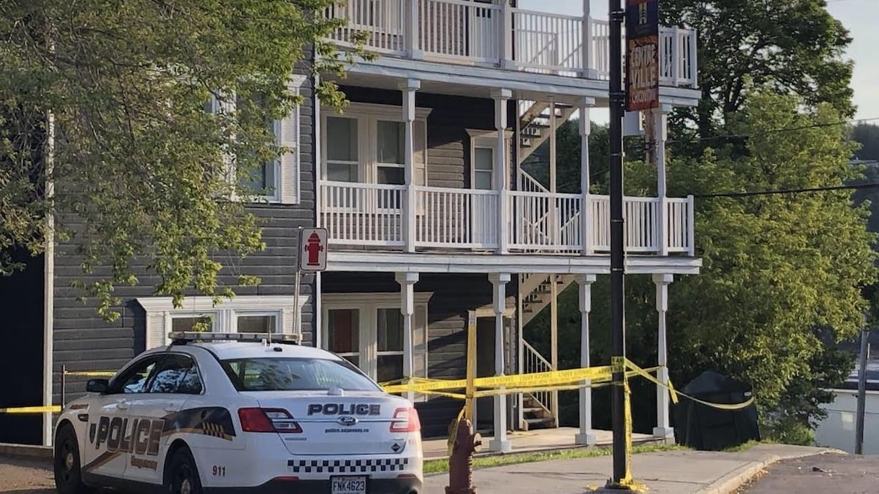 Une voiture de patrouille stationnée devant un immeuble à logements entouré d'un périmètre de sécurité.