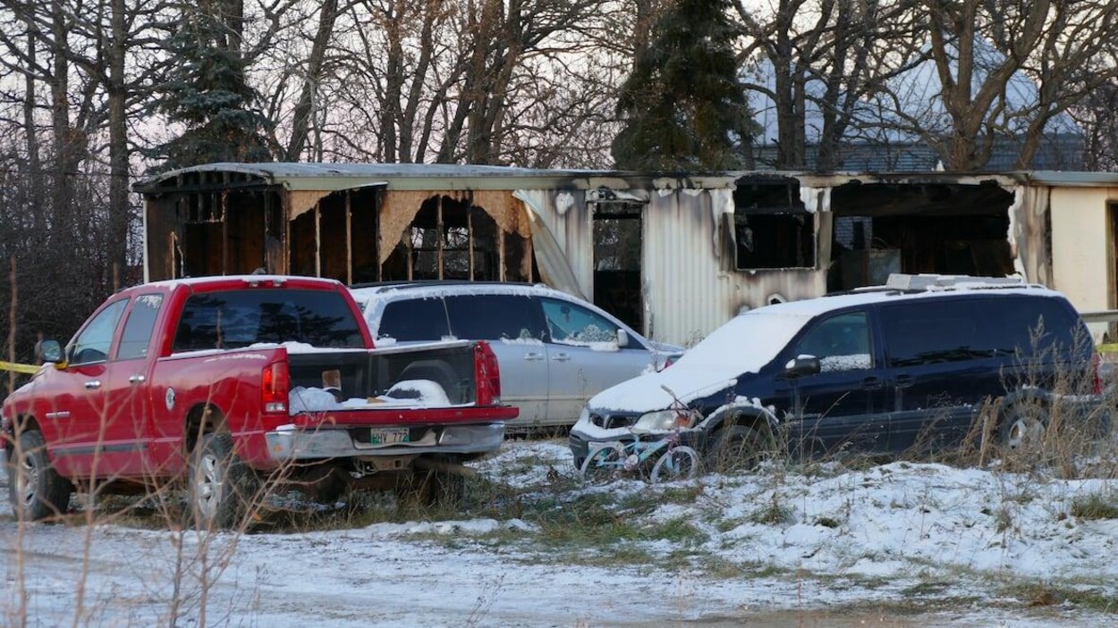 Un camion de style pick-up et deux camionnettes sont stationnés devant la structure d'une maison modulaire incendiée.