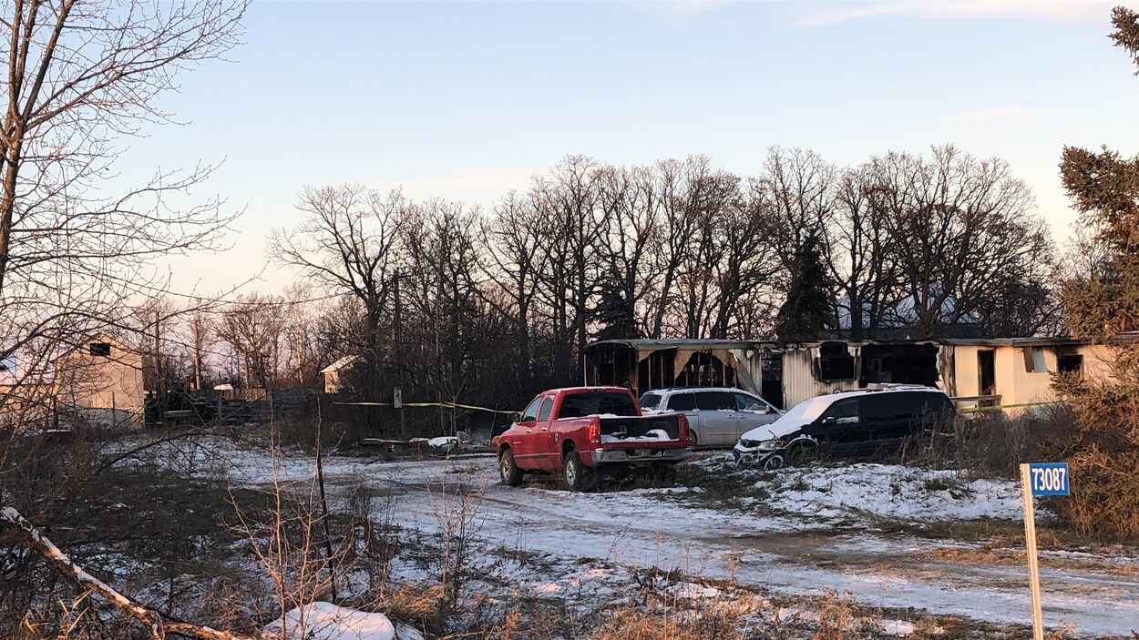 Un camion de style pick-up et deux camionnettes stationnées devant la structure d'une maison mobile incendiée.