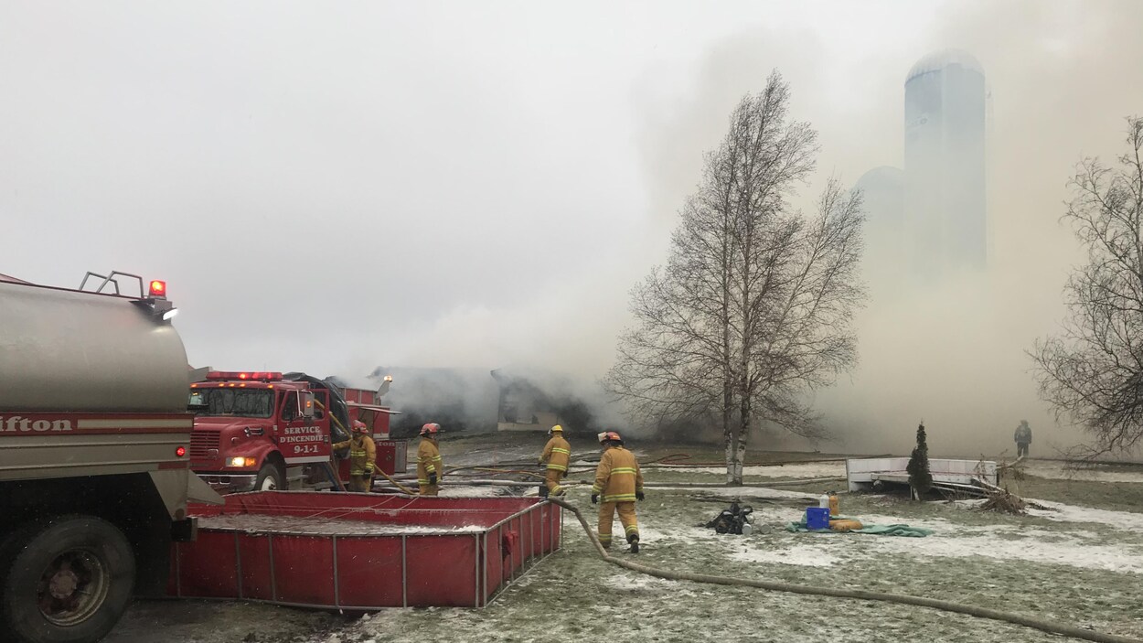 Plus d'une vingtaine de pompiers ont été appelés à combattre l'incendie sur le rang 5, à Saint-Malo. On voit ici les pompiers puiser de l'eau d'un bassin. Il y a énormément de fumée derrière eux.