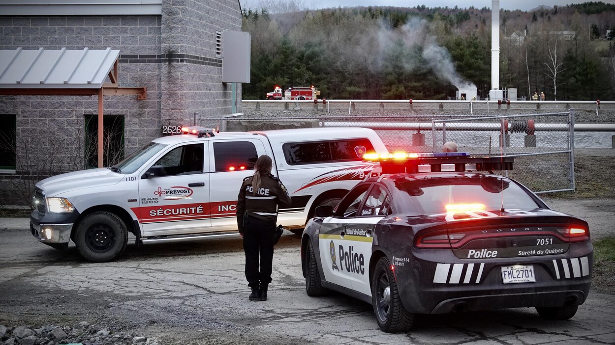 Une policière près de son autopatrouille observe des pompiers qui s'affairent autour d'une tour de télécommunication. Une fumée blanche sort d'une cabane située tout près de la tour.