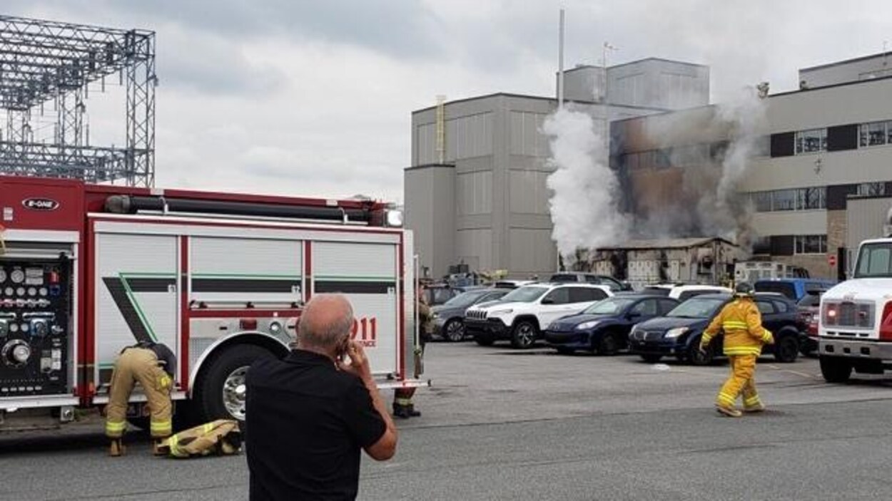 Homme au téléphone devant un camion de pompier et une usine où on observe des colonnes de fumées qui sortent de la bâtisse,