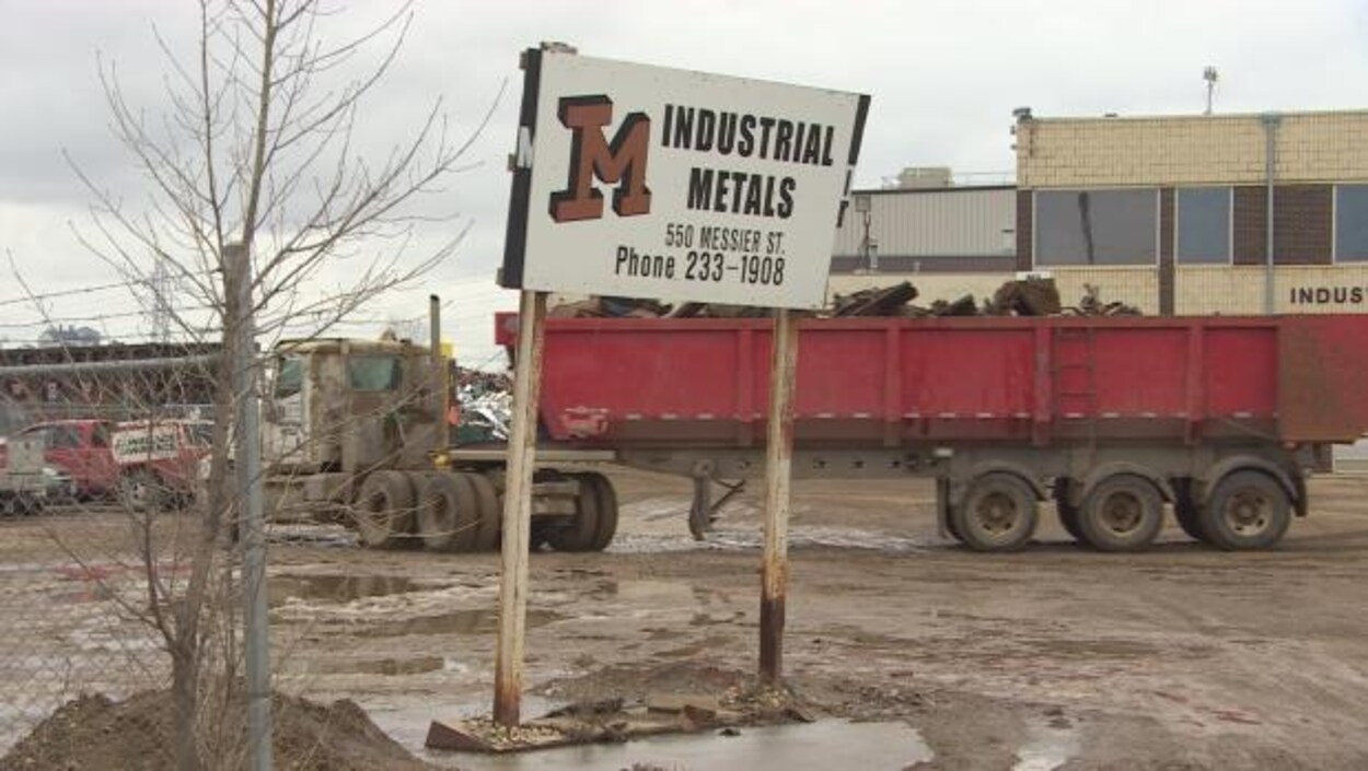 L'avant-plan de la photo montre une affiche de l'usine avec son logo et son adresse. À l'arrière, on voit le bâtiment et un camion transportant des déchets métalliques.