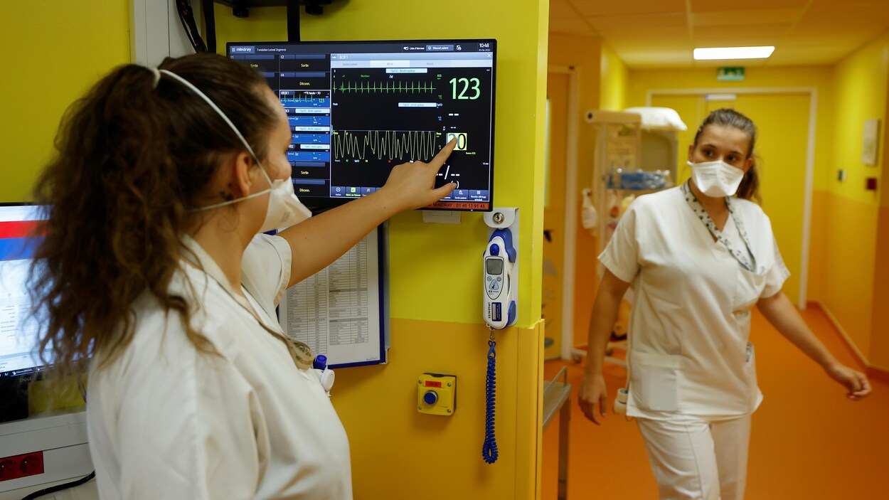 Une infirmière pose l'index sur l'écran tactile accroché au mur de l'hôpital, sur lequel on voit des chiffres et des graphiques, pendant qu'une autre infirmière entre dans la pièce.