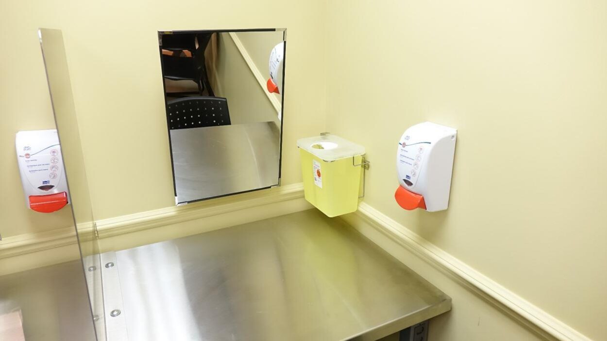 Le cubicule comporte un miroir, une table d'acier, un réservoir de liquide désinfectant et une poubelle pour seringues usagées.