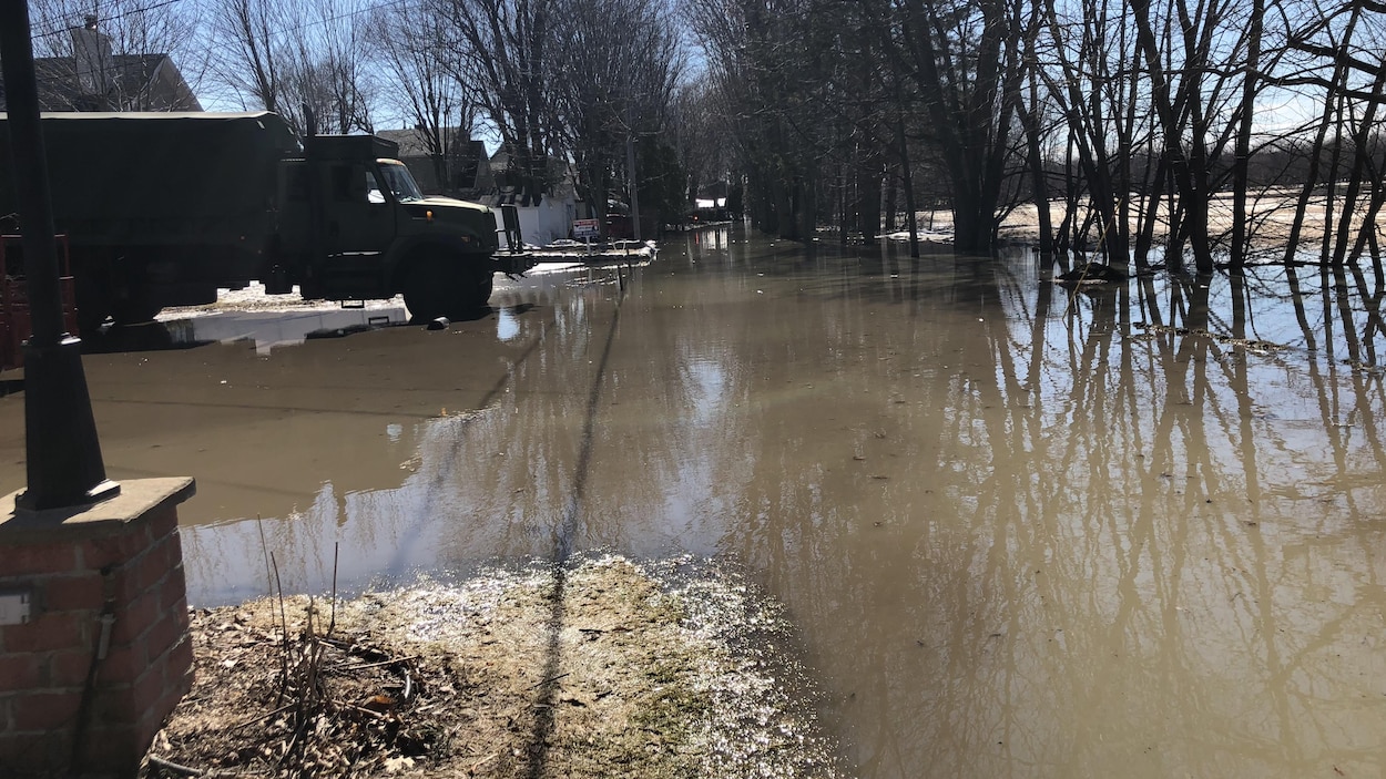 Dans un secteur rural, de l'Eau inonde ce qui était une rue. Un camion de l'armée est stationné devant des maisons.
