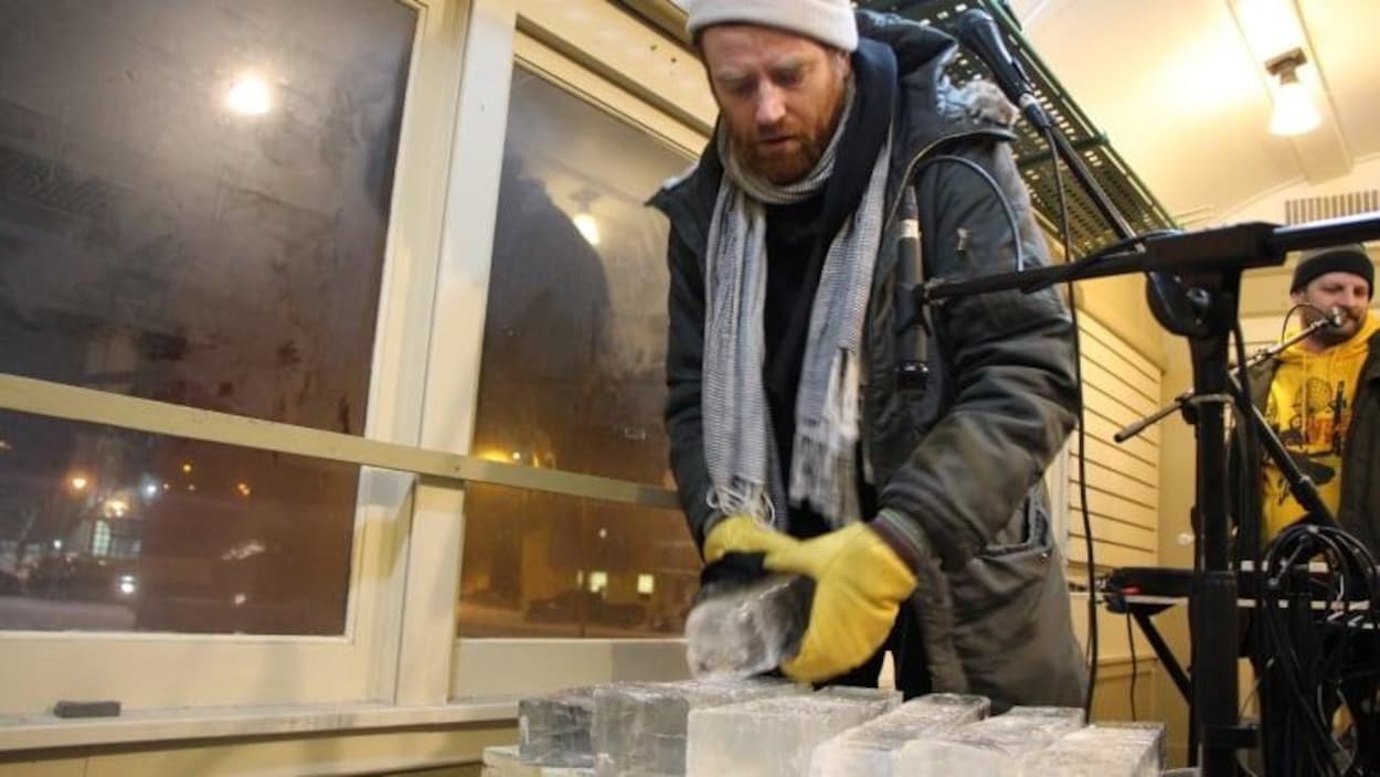 Un homme debout qui tient un morceau de glace. Près de lui des micros et d’autres instruments.
