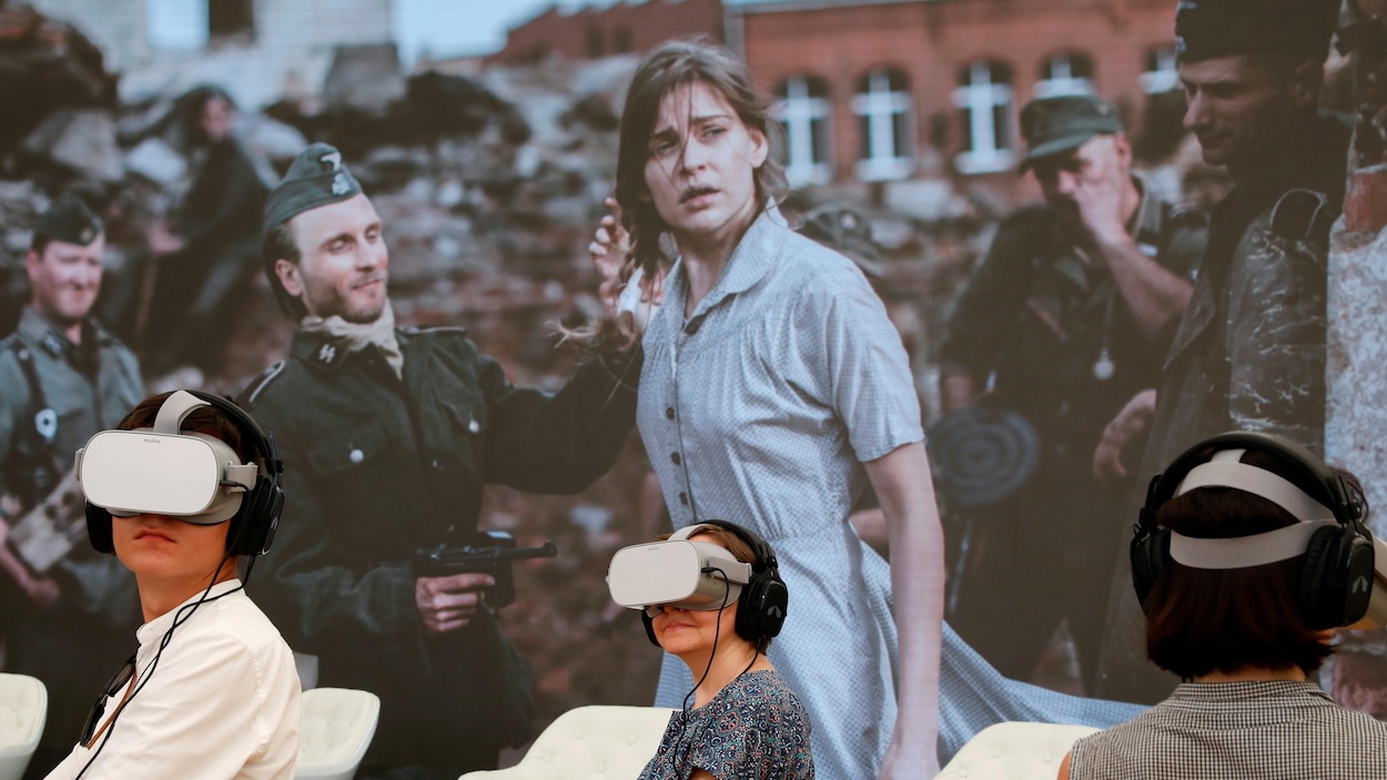 Des personnes portent des casques de réalité virtuelle et des écouteurs devant une murale montrant une jeune femme apeurée entourée de soldats nazis.