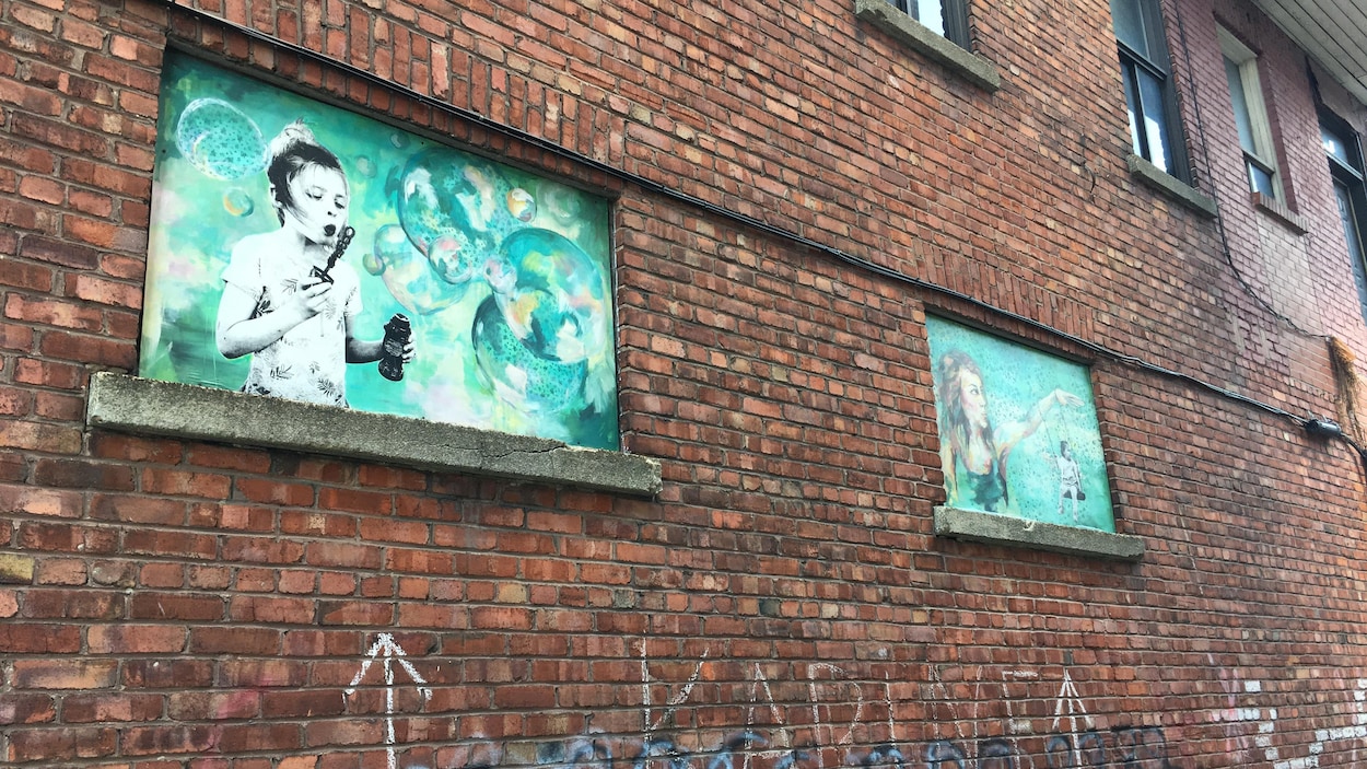 Deux toiles où des enfants s'amusent ornent les fenêtres d'une bâtisse dans une ruelle.