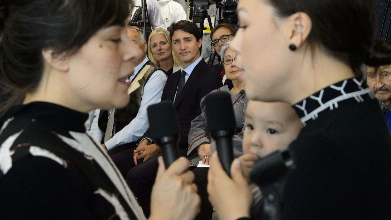 Deux femmes chantent devant Justin Trudeau assis sur une chaise.