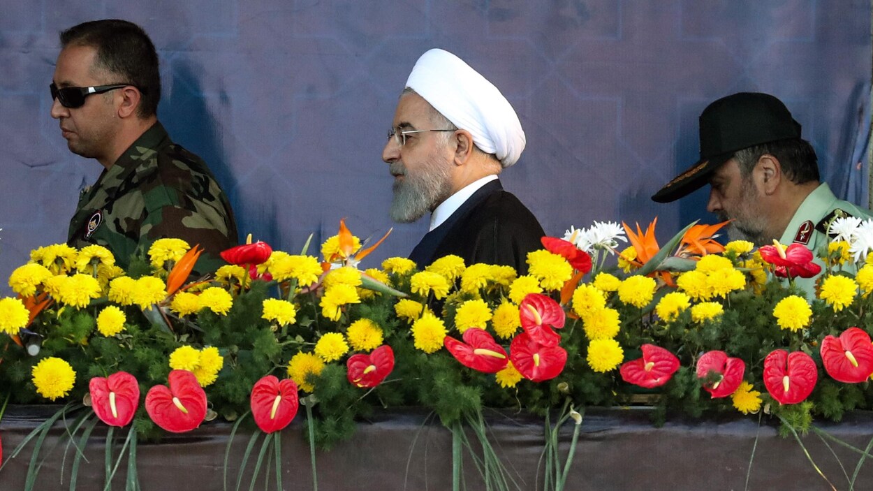 Un homme portant la barbe et un turban blanc marche derrière une haie de fleurs entre deux militaires.