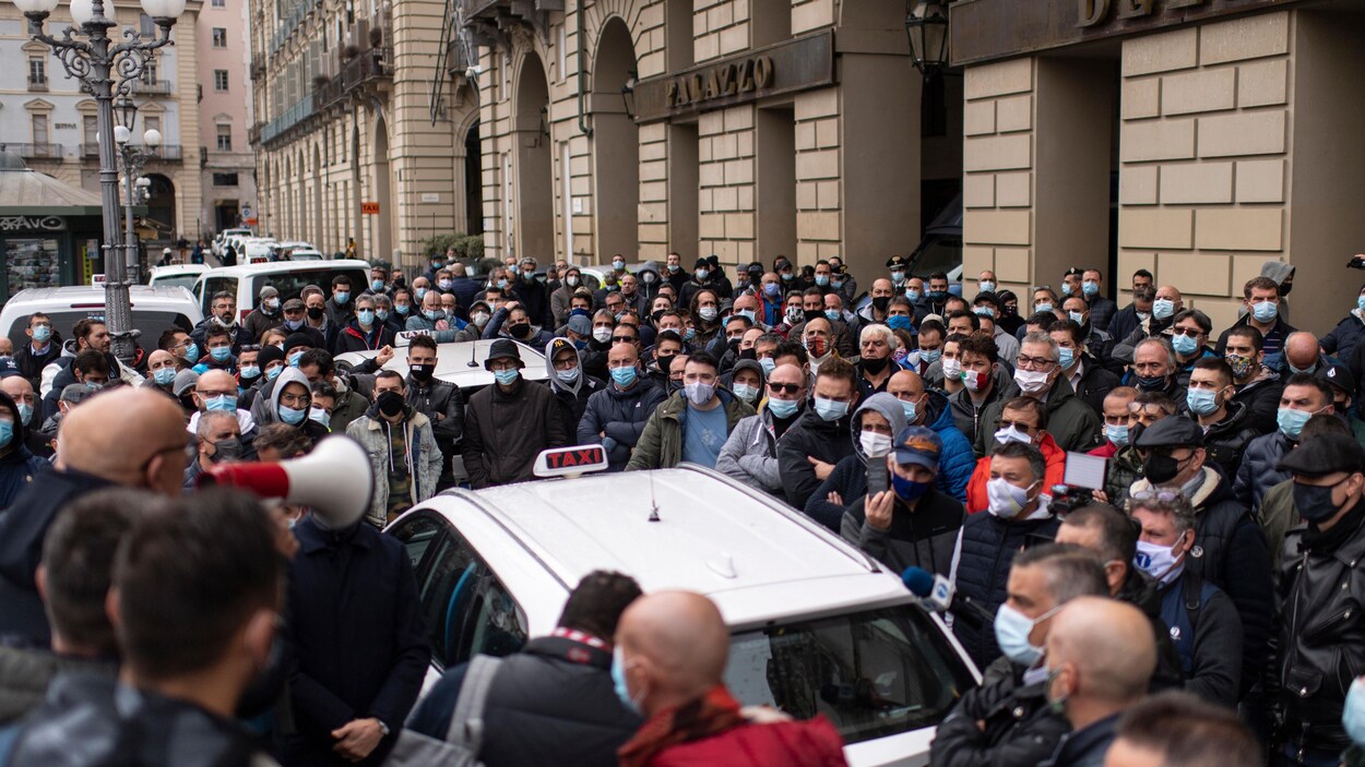 Des dizaines de personnes masquées sont rassemblées sans grande distanciation physique autour de taxis. Un homme s'adresse à eux avec un mégaphone.