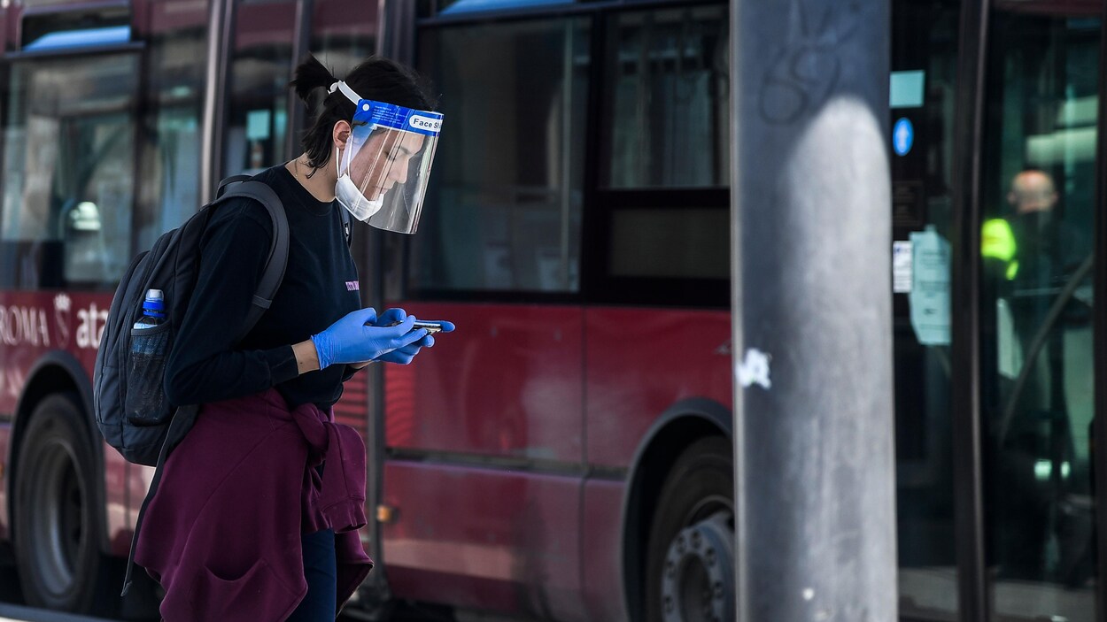 Une Italienne portant une visière et un masque consulte son téléphone cellulaire près d'un autobus.