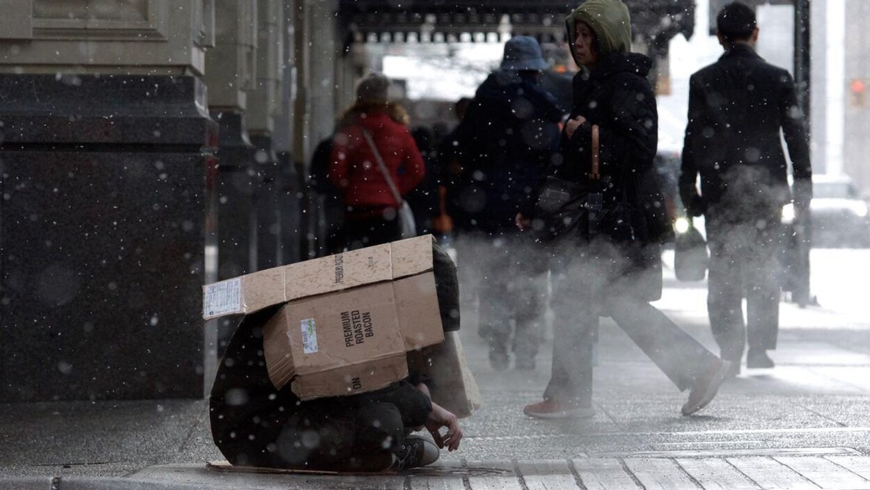 Un sans-abri assis dans le rue, avec des passants en arrière-plan