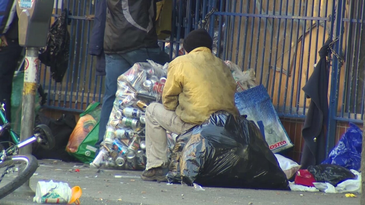 Un homme assis sur un sac poubelle contenant des canettes, d'autres sacs l'entourent sur le trottoir.