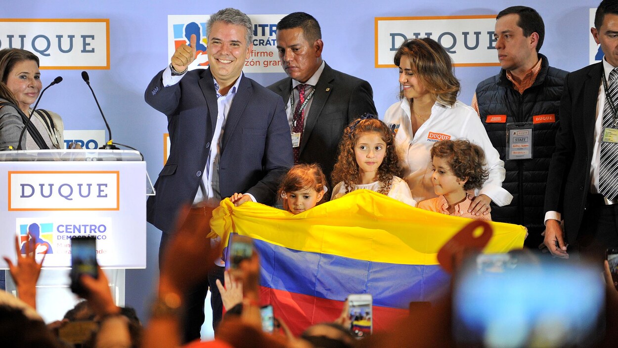Ivan Duque salue ses partisans sur une estrade, en plaçant un pouce en l'air. Il est notamment accompagné d'une femme et de trois enfants qui tiennent un drapeau colombien.