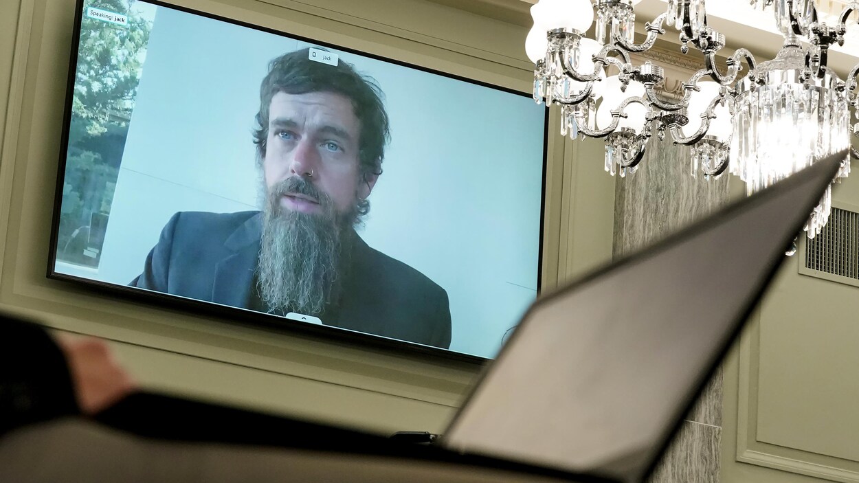 Le visage de l'homme est visible sur un écran géant dans une salle décorée élégamment, où on voit un chandelier.