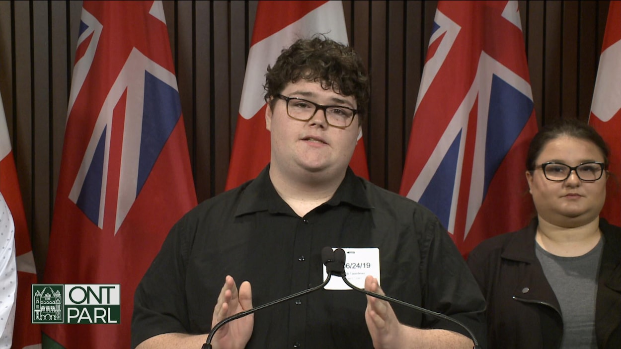 Un étudiant gesticule avec ses mains en parlant à un podium avec des drapeaux de l'Ontario derrière lui.