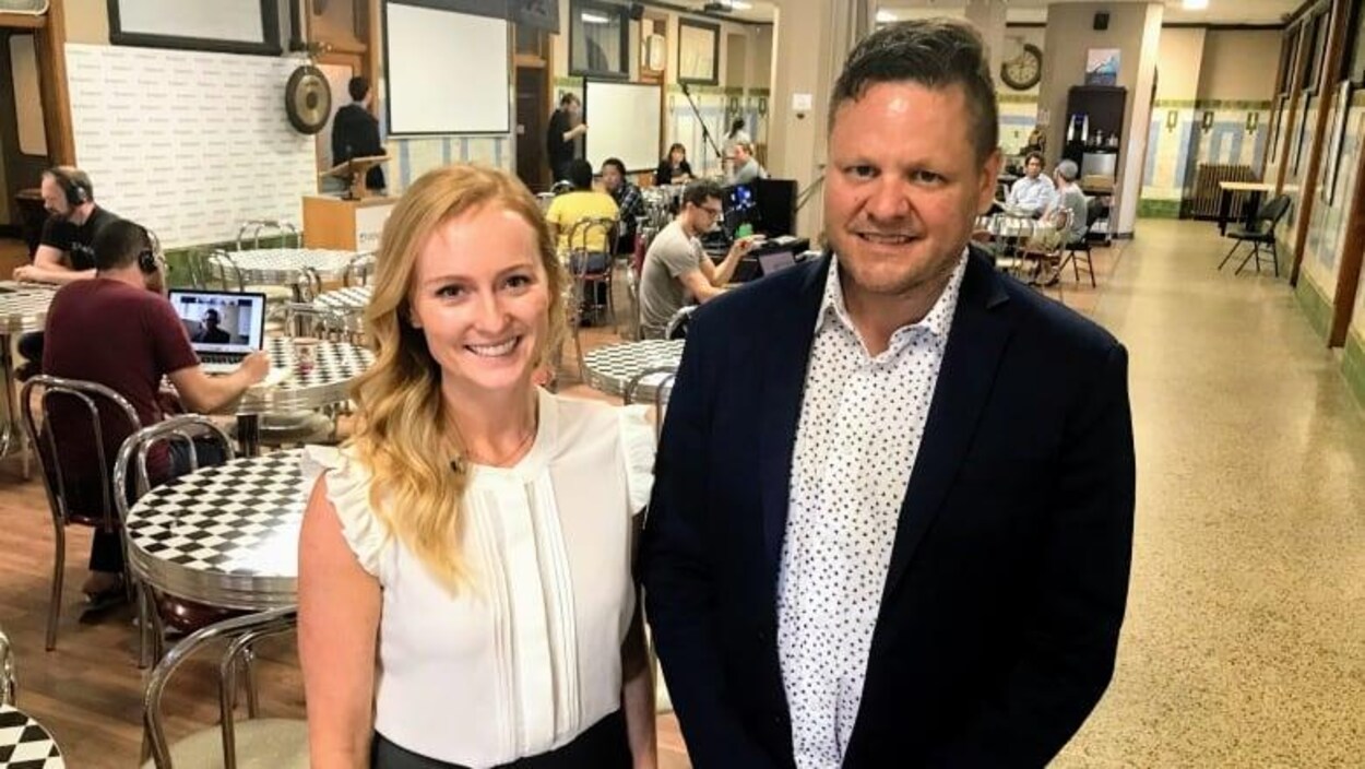 Jacqueline Cook et Brendan King se tiennent debout dans une salle de travail dans l’entreprise qui ressemble à une cafétéria.