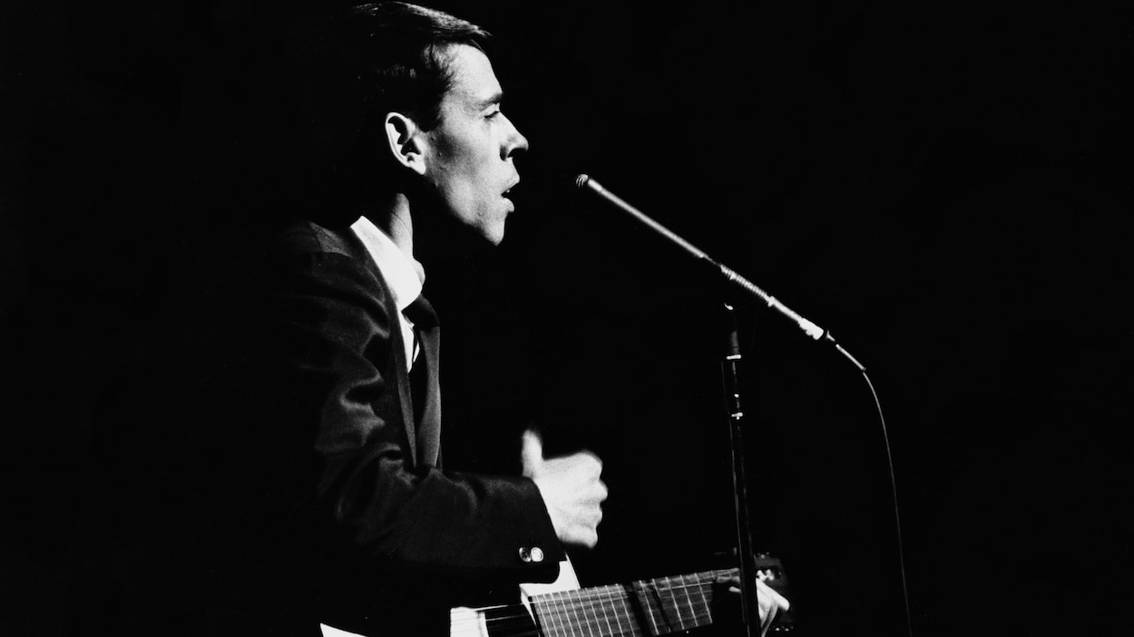 Sur une scène, Jacques Brel donne une performance s'accompagnant de sa guitare, un micro sur pied devant lui.