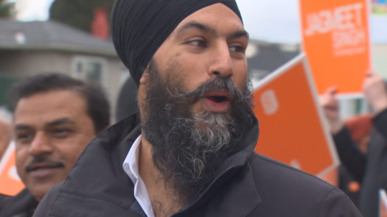 Gros plan sur Jagmeet Singh avec des gens qui tiennent des pancartes oranges à l'arrière.