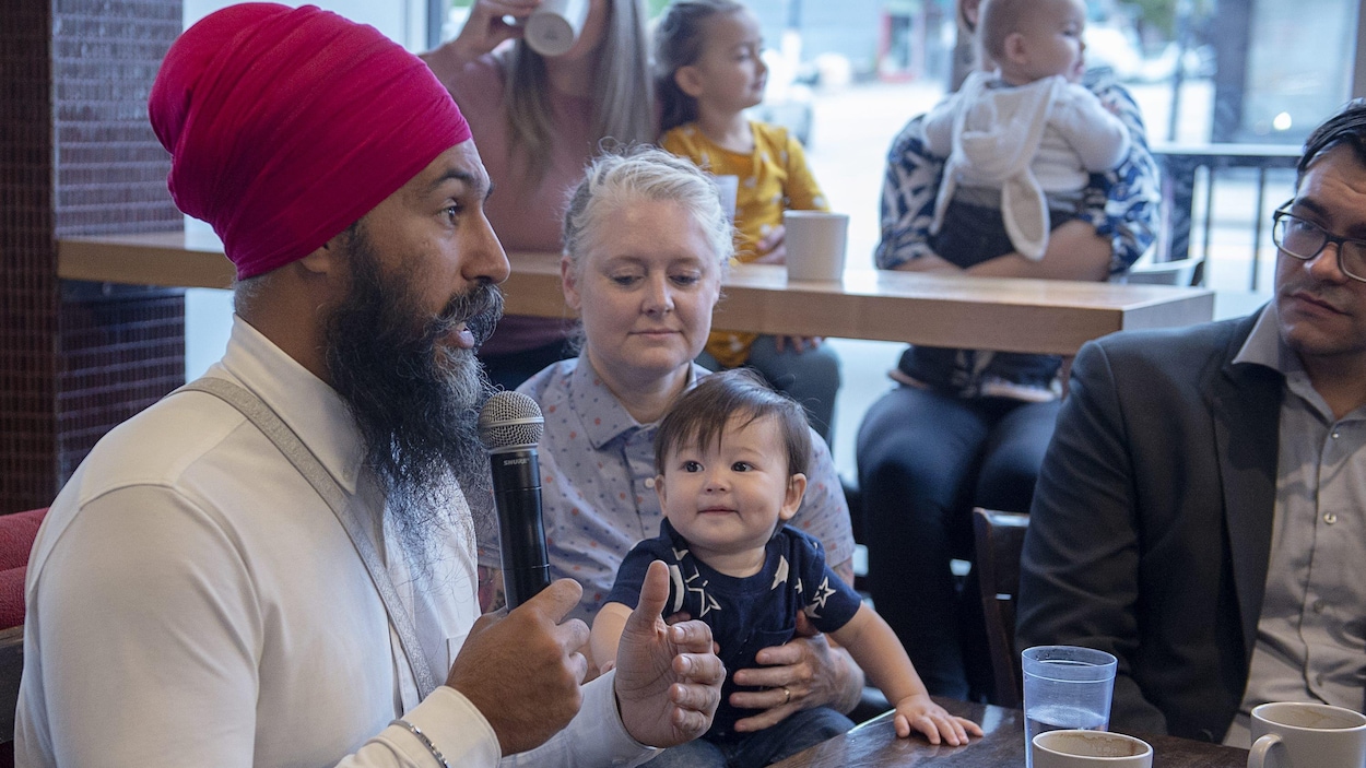Jagmeet Singh parle dans un micro, assis à une table à côté d'une femme qui tient un bébé sur ses genoux.