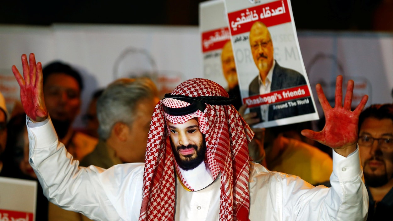 Un homme portant un masque de Mohammed ben Salmane lève ses deux mains peintes en rouge. Derrière lui, des gens brandissent des photos de Jamal Khashoggi.