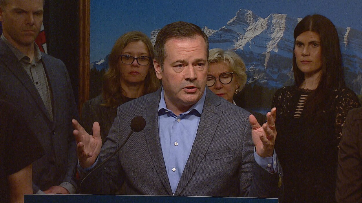 Jason Kenney parle devant un podium. Des gens sont debout derrière lui.