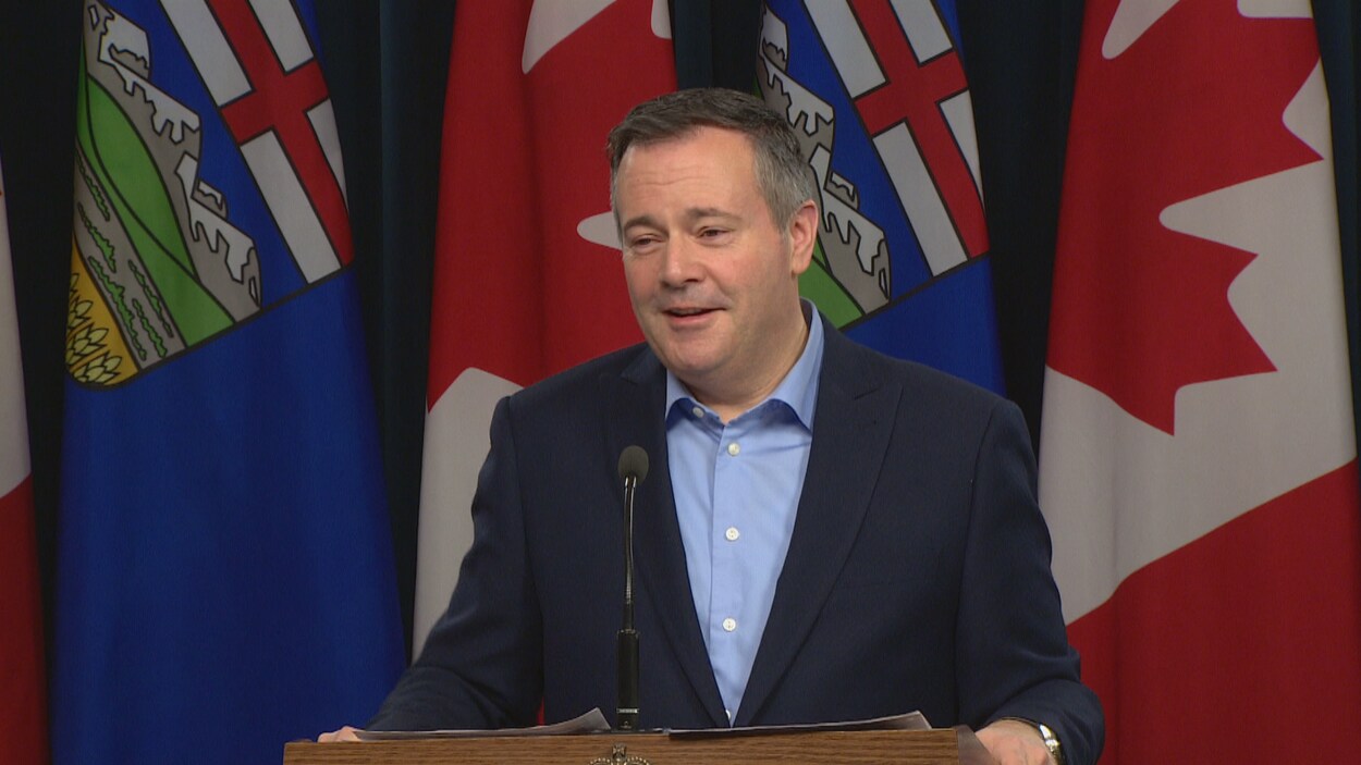 Plan moyen de Jason Kenney parlant lors d'une conférence de presse. Il est debout devant des drapeaux canadiens et albertains.